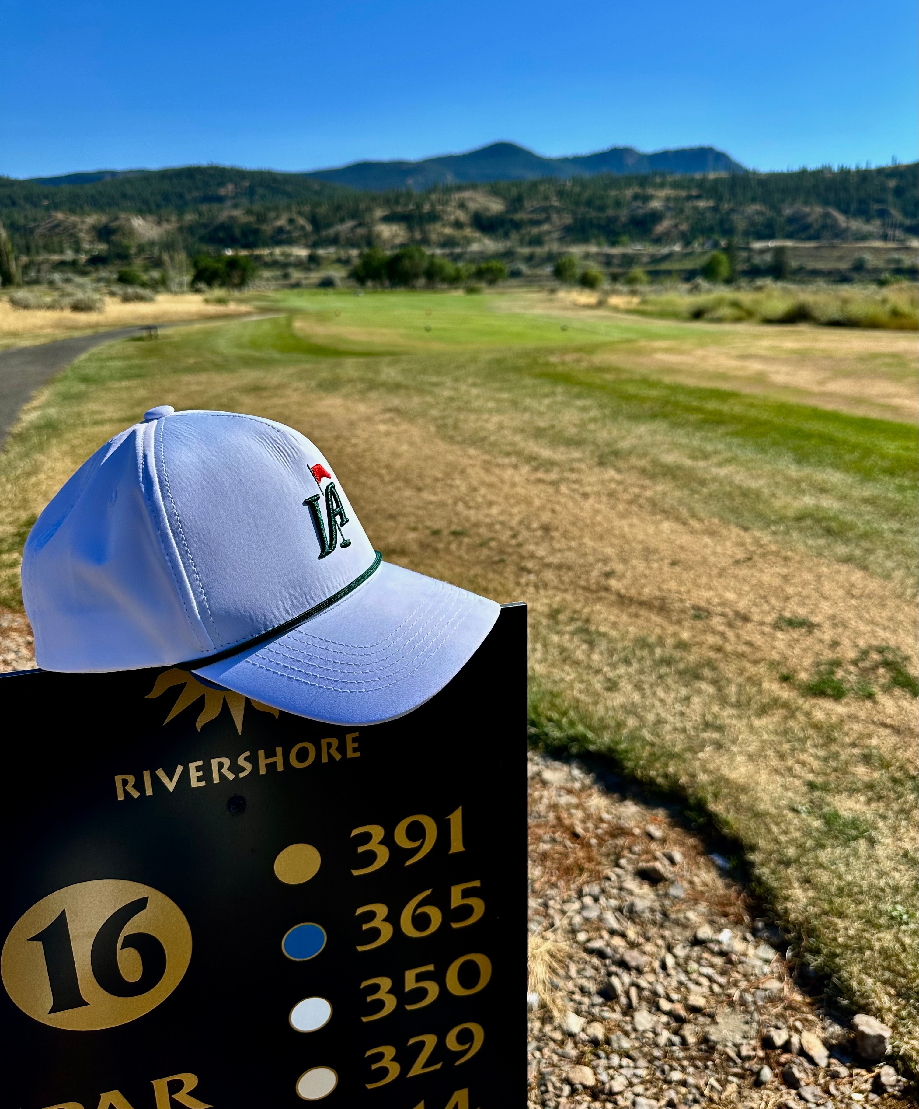 A white golf hat rests on a sign indicating the 16th hole at Rivershore Golf Course, with a view of distant mountains and a sunny blue sky in the background.